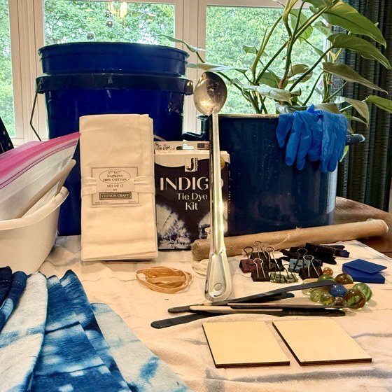 Workshop supplies laid out on a table: cotton napkins, indigo kit, bucket, gloves, rubber bands, clamps, marbles and a wooden dowel