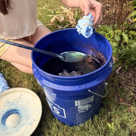 A gloved hand lowering a bound napkin into an outdoor indigo vat, with a soak pot beside it